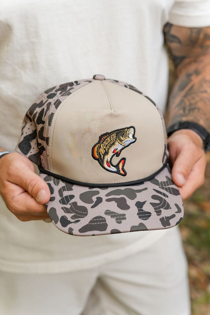 A person with arm tattoos wearing a white shirt holds the Mucker Bucket Big Bass Daddy Duck Camo Bucket hat, which features beige and gray camouflage, a breathable water-resistant finish, and an embroidered largemouth bass patch.