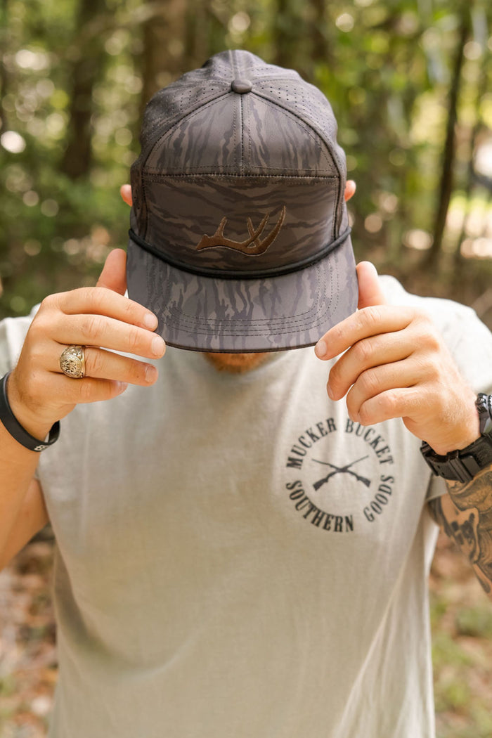 A person in a gray T-shirt and a Mucker Bucket Buck Season Lowcountry Camo Bucket hat adjusts the brim outdoors, face hidden. The T-shirt reads Wicker Bucket Southern Goods.