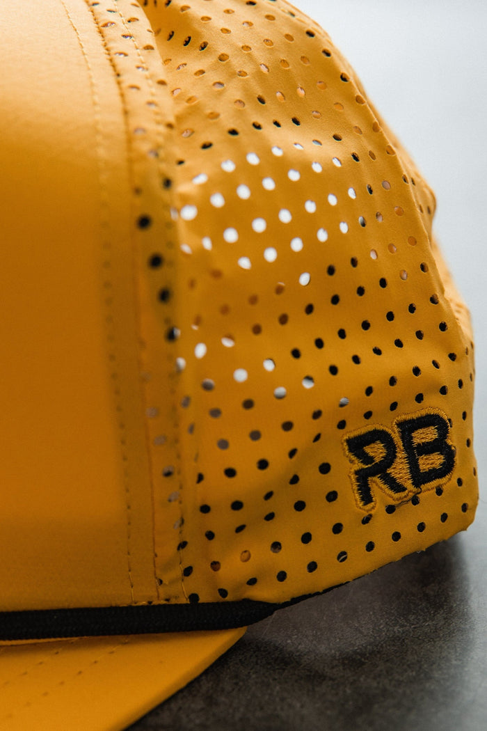 Close-up of the Burn The Ships - Gold Snapback by Relentless Betrayal, showcasing its yellow color with black perforated dots and RB embroidery. The snapback enclosure and textured details stand out against a blurred background.