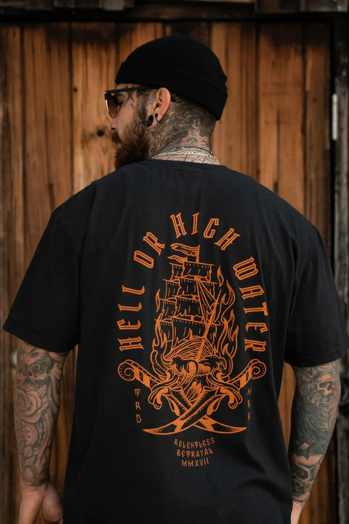 A tattooed man wearing a beanie and the Hell or High Water Premium Tee by Relentless Betrayal, showcasing an orange ship design on a black T-shirt, exudes unwavering spirit. The backdrop is framed with wooden panels.