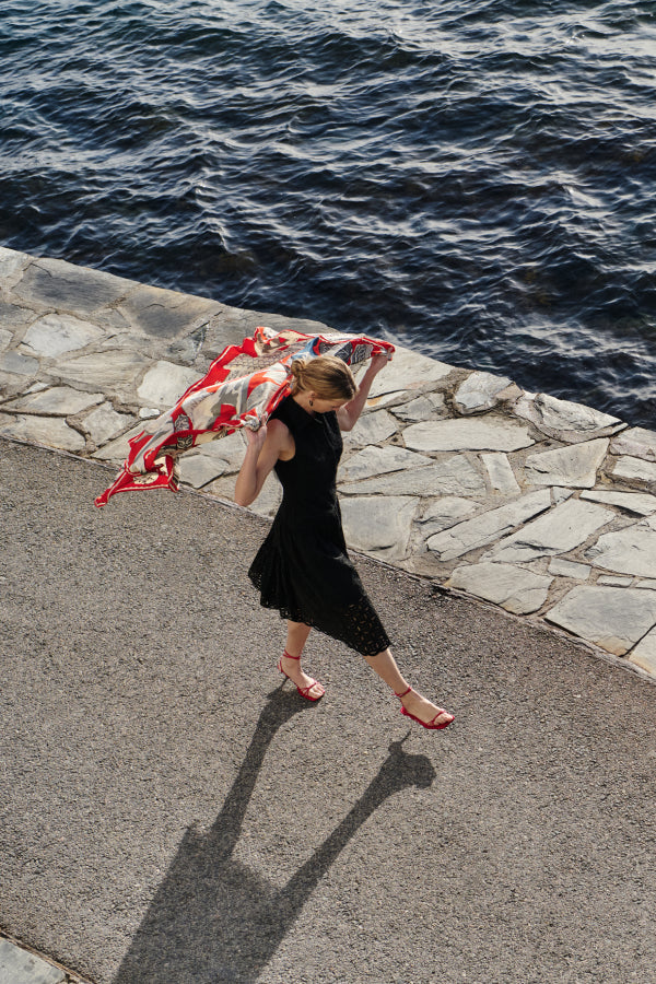 Woman in a black dress holding a red and white patterned scarf by the water. | Exclude