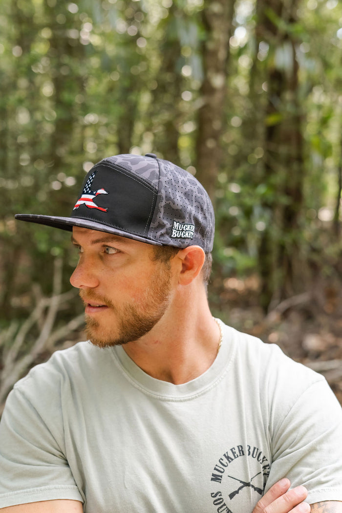 A bearded man wearing a Mucker Bucket "Merica Duck Blackout Duck Camo" bucket hat with an embroidered patch stands outdoors in a forest, looking to his left amid green trees and foliage.