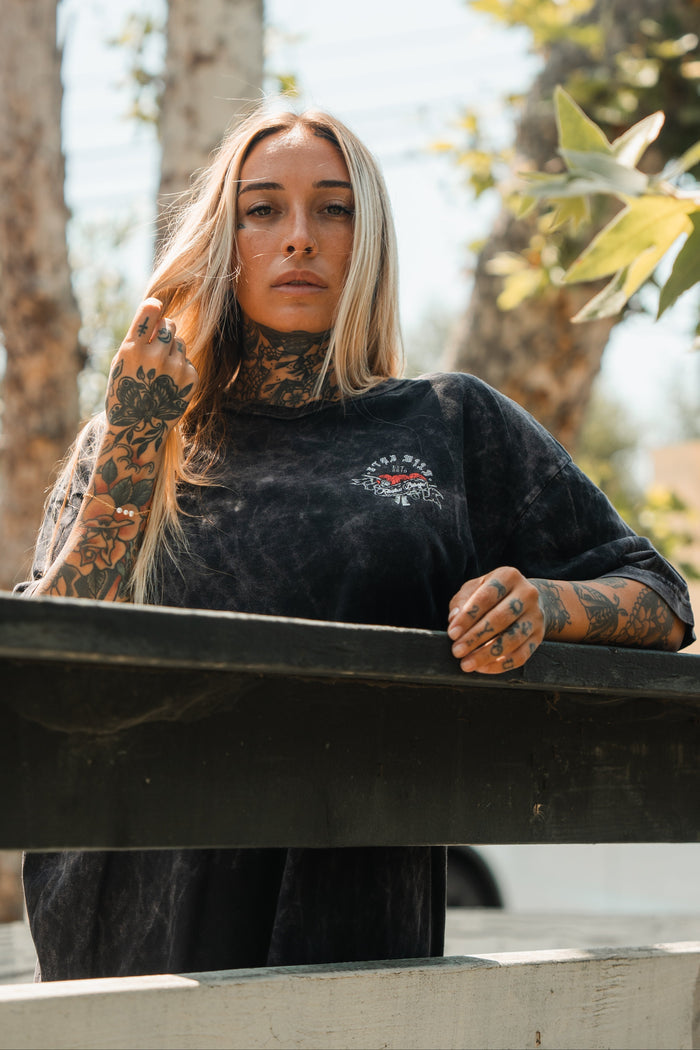 A woman with long blonde hair and tattoos stands outdoors in sunlight, wearing the Relentless Betrayal Stay Wild Premium T-Shirt - Stone, holding her hair as light filters through the trees.