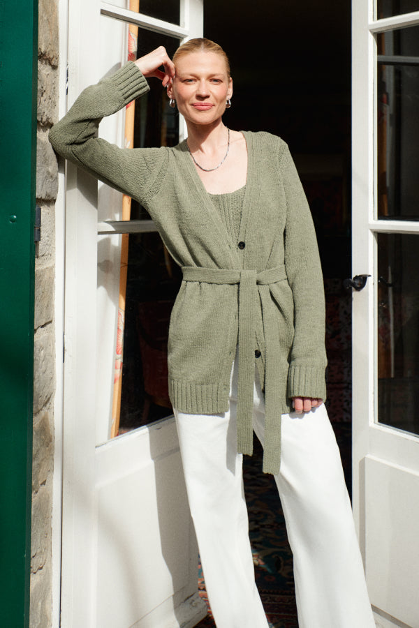 Woman wearing a green cardigan and white pants standing in front of a door. | Exclude | Grid