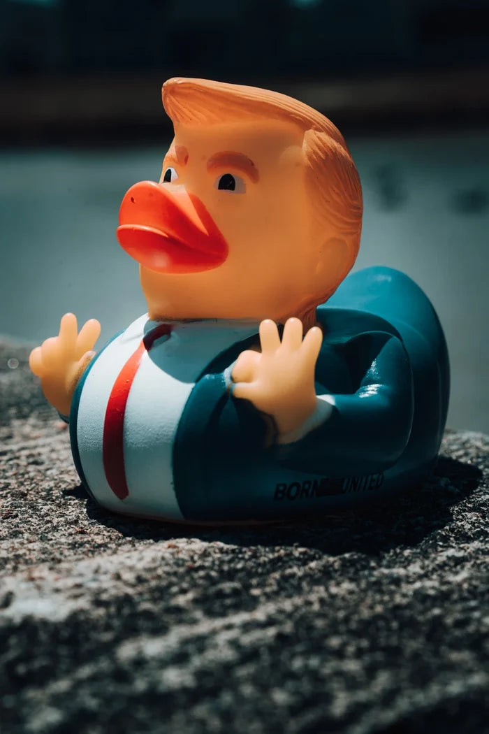 Product #47 by Born United Private Label— a rubber duck in a blue suit and red tie with orange hair and lips, rests on stone. Sunlight casts shadows, accentuating its playful design as a nod to the latest trend. #IYKYK.
