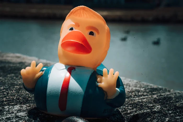 A #47 rubber duck by Born United Private Label wears a business suit with an orange tie on a concrete surface near water, capturing the perfect #trend moment. Its human-like features and blurred background create an IYKYK scene for fans of quirky elegance.