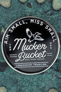 A round black sticker with white text reads Mucker Bucket Southern Goods, featuring an illustration of a duck above it, evocative of the Aim Small, Miss Small Lucky Huntin Coin. The background showcases star-like patterns on a textured greenish-blue surface.