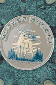 The Chasin Tail Lucky Huntin Coin by Mucker Bucket features a stag in mountains and trees. With Crown Tail and Made in the Netherlands on it, set against a speckled green and brown background, its perfect for your next adventure.