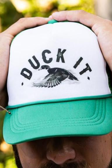 A person wears the Duck It Green Trucker Hat by Mucker Bucket, featuring a white and green design with DUCK IT and a flying duck in high-quality screen print. Their hands, decorated with bracelets and a watch, rest atop the cap against a backdrop of blurred greenery.