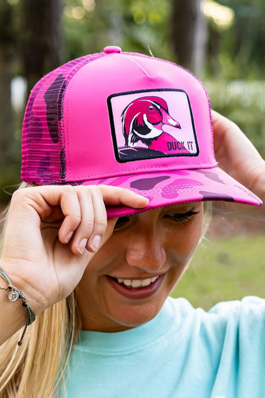 A smiling woman adjusts a Mucker Bucket "Duck It" Pink Camo Trucker Bucket hat with a wood duck patch. She stands outdoors, green foliage softly blurred behind her.