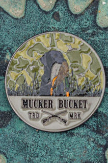 The Got One Lucky Huntin Coin is a round metal pin with a person shoveling, crossed shovels, and Mucker Bucket TRD MRK on it. Its green camouflage background hints at adventure, making it a lucky charm for any bold collector.
