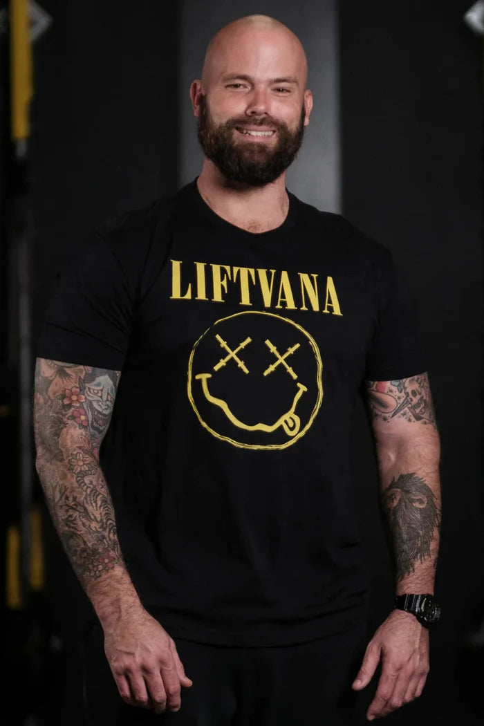 A smiling, bald, bearded man with tattooed arms stands by gym equipment, wearing the "Liftvana" Men's Tee from Tango Charlie Apparel—showcasing a black shirt with a yellow smiley face and X eyes for true lifter spirit and grunge style.
