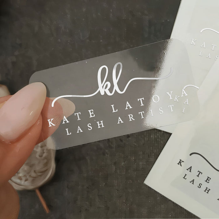 Transparent clear vinyl branding sticker with silver foil script initials and serif text, glossy finish shown on hand for close-up detail – XOXOKristen.