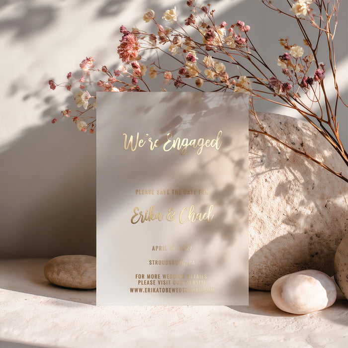 A gold foil vellum Save the Date card placed among dried flowers and soft shadows. The natural lighting highlights the transparent material, creating a dreamy, romantic atmosphere perfect for luxury wedding stationery - XOXOKristen