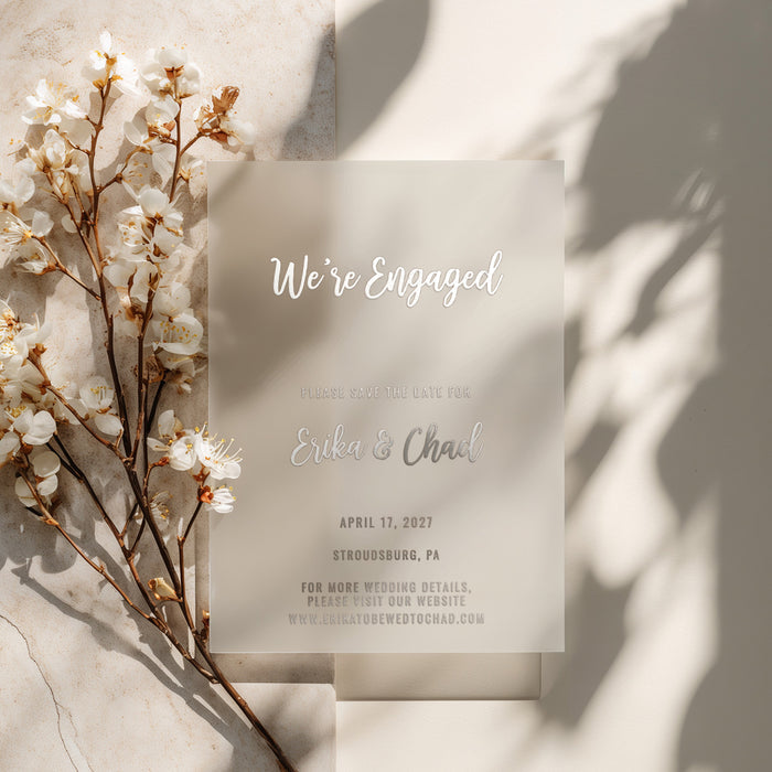 A delicate silver foil vellum card placed beside white blooming flowers on a warm-toned background. The light passes softly through the vellum, highlighting the text “We’re Engaged” — ideal for romantic, airy wedding stationery photography - XOXOKristen