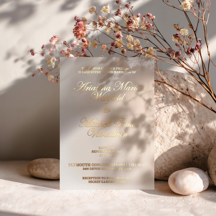 vellum wedding invitation printed in luxurious gold foil lettering, standing upright among soft dried flowers and warm light, highlighting its delicate transparency and elegant typography – XOXOKristen